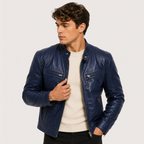 A man wearing a dark blue leather jacket over a cream ribbed sweater and black pants, photographed against a light gray studio background.