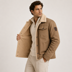 A man wearing a tan sherpa-lined jacket over a cream zip-up sweater and light beige trousers, standing against a plain light gray studio background.