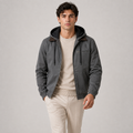 A man wearing a dark gray hooded jacket with brown collar details, layered over a beige crewneck sweater and cream pants, standing against a neutral gray studio background.