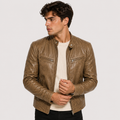 A man wearing a brown faux-leather biker jacket over a cream ribbed sweater and black pants, photographed against a light gray studio background.