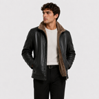 A man wearing a black leather jacket with a brown shearling-style collar, layered over a white ribbed sweater and black trousers, photographed against a light gray studio background.