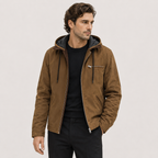 A man wearing a brown hooded jacket with a dark gray interior lining, styled over a black t-shirt and black pants, photographed against a light gray studio background.