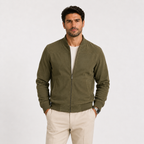A man wearing an olive green zip-up bomber jacket over a white t-shirt and beige trousers, standing against a light gray studio background.