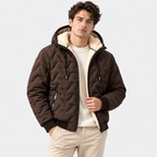 A man wearing a dark brown quilted puffer jacket with a cream sherpa-lined hood and collar, styled over a cream knit sweater and beige trousers, against a light gray studio background.