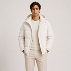 A man wearing a cream-colored hooded puffer jacket over a matching cream sweater and light beige trousers, standing against a plain light gray studio background.