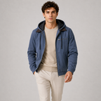 A man wearing a blue hooded zip-up jacket with dark brown collar lining, layered over a beige crewneck sweater and cream trousers, standing against a neutral gray studio background.