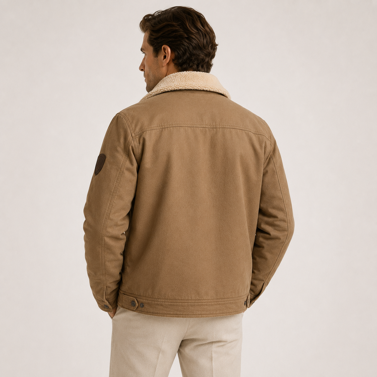 A man shown from the back wearing a tan canvas jacket with a cream sherpa collar, paired with light beige trousers, against a plain light gray studio background.