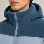 Close-up of a person wearing a blue corduroy hooded puffer jacket with a light gray lower panel and zipper.