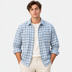 A man wearing a light blue and white plaid button-down shirt over a cream t-shirt and beige pants, standing against a plain white studio background.