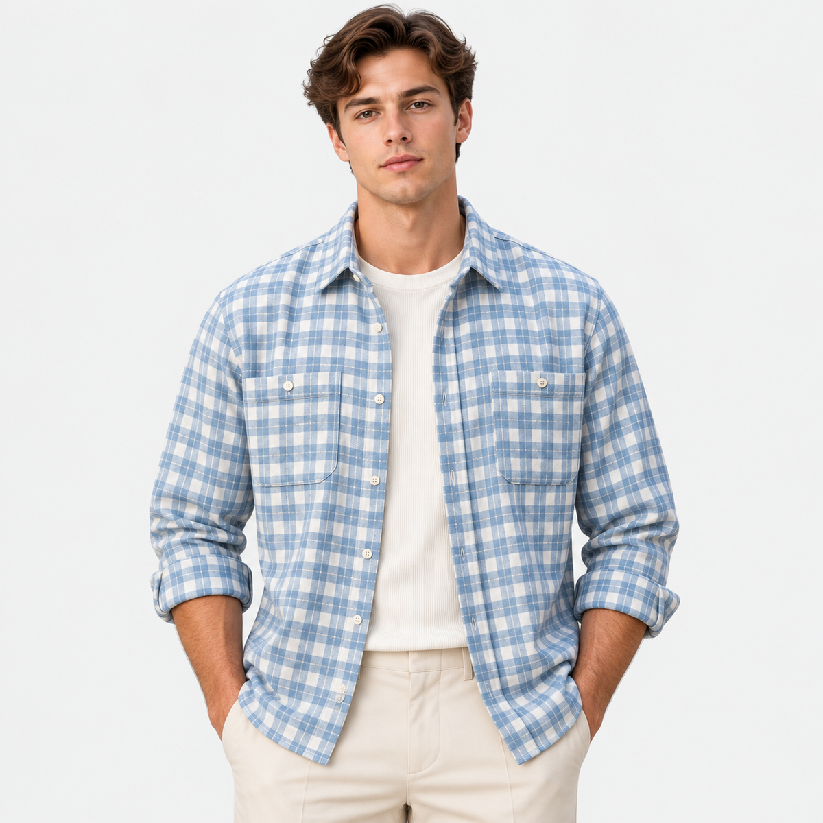 A man wearing a light blue and white plaid button-down shirt over a cream t-shirt and beige pants, standing against a plain white studio background.