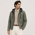 A man wearing an olive green hooded jacket over a beige sweater and white trousers, photographed against a plain light gray studio background.