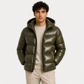 A man wearing a shiny olive green hooded puffer jacket over a cream ribbed sweater and beige trousers, photographed against a plain light gray studio background.