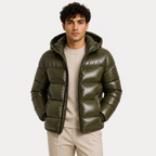 A man wearing a shiny olive green hooded puffer jacket over a cream ribbed sweater and beige trousers, photographed against a plain light gray studio background.