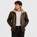 A man wearing a brown leather aviator-style jacket with a beige sherpa collar, layered over a white ribbed sweater and black trousers, photographed against a light gray studio background.