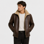 A man wearing a brown leather aviator-style jacket with a beige sherpa collar, layered over a white ribbed sweater and black trousers, photographed against a light gray studio background.