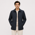 A man wearing a dark navy blue diamond-quilted jacket over a light beige crewneck sweater and beige trousers, standing against a solid off-white background.