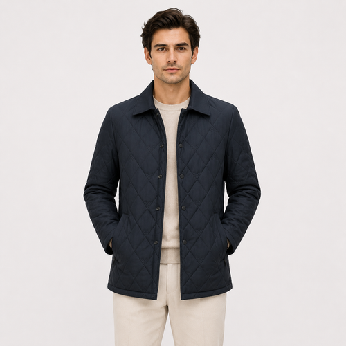 A man wearing a dark navy blue diamond-quilted jacket over a light beige crewneck sweater and beige trousers, standing against a solid off-white background.