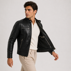 A man wearing a black leather moto jacket over a cream polo shirt and beige trousers, photographed against a light gray studio background.