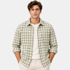A man wearing a light sage green and white gingham plaid button-down shirt over a white t-shirt and cream pants, standing against a plain white studio background.