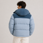 A man viewed from the back wearing a light blue and dark blue color-blocked puffer jacket with a hood, paired with off-white pants, against a plain light gray studio background.
