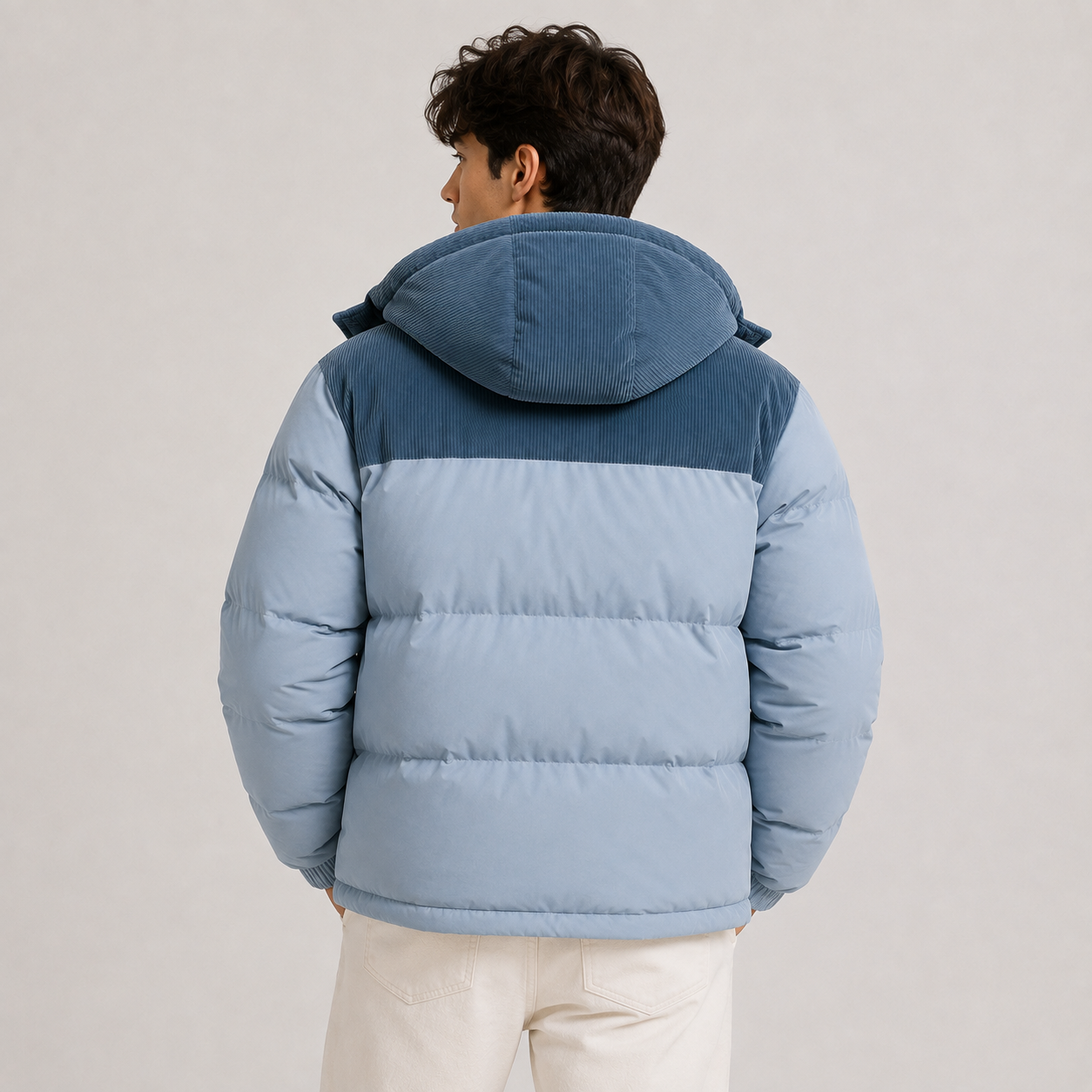 A man viewed from the back wearing a light blue and dark blue color-blocked puffer jacket with a hood, paired with off-white pants, against a plain light gray studio background.