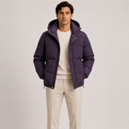 A man wearing a dark purple hooded puffer jacket over a cream sweater and beige trousers, standing against a light gray studio background.