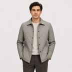 A man wearing a light gray diamond-quilted button-up jacket over a cream crewneck sweater and dark gray trousers, standing against a light gray studio background.