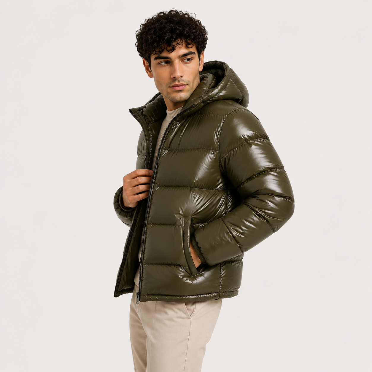 A man wearing a shiny olive green hooded puffer jacket over a light beige shirt and beige pants, photographed against a plain light gray studio background.