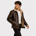 A man wearing a brown leather aviator-style jacket with a beige sherpa lining, paired with a white t-shirt and black pants, photographed against a solid light gray background.