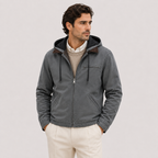 A man wearing a dark gray zip-up hooded jacket over a beige sweater and white pants, standing against a light gray studio background.