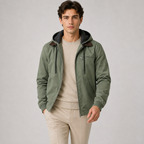 A man wearing an olive green hooded jacket with dark brown collar lining, a beige crewneck sweater, and beige trousers, standing against a light gray studio background.