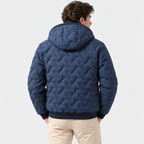 A man viewed from the back wearing a navy blue quilted hooded jacket with a geometric chevron pattern, paired with beige pants, against a plain light gray studio background.