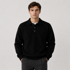 A man wearing a black knit polo sweater over a white undershirt and dark gray trousers, standing against a light gray studio background.