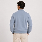 A man viewed from behind wearing a light blue knit quarter-zip sweater and beige trousers, standing against a plain light gray studio background.