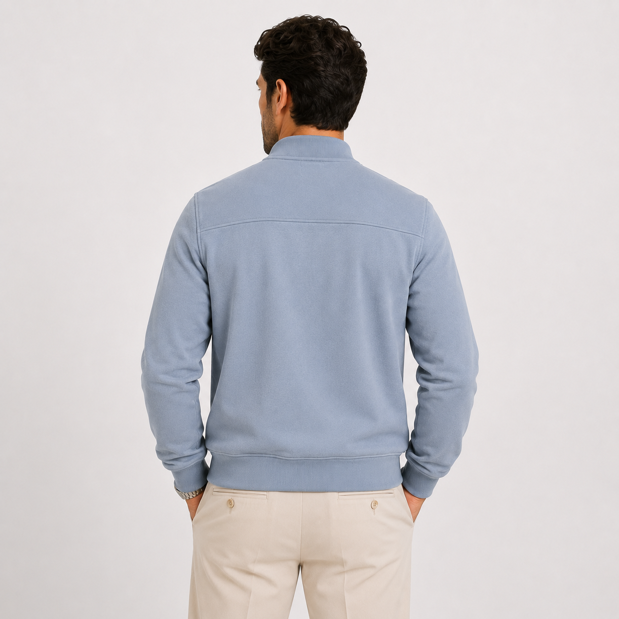 A man viewed from behind wearing a light blue knit quarter-zip sweater and beige trousers, standing against a plain light gray studio background.