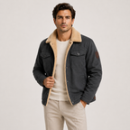 A man wearing a dark charcoal gray jacket with a beige sherpa collar, layered over a cream t-shirt and beige trousers, photographed against a light gray studio background.