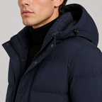 A close-up of a man wearing a navy blue hooded puffer jacket over a black turtleneck sweater, photographed against a light gray studio background.