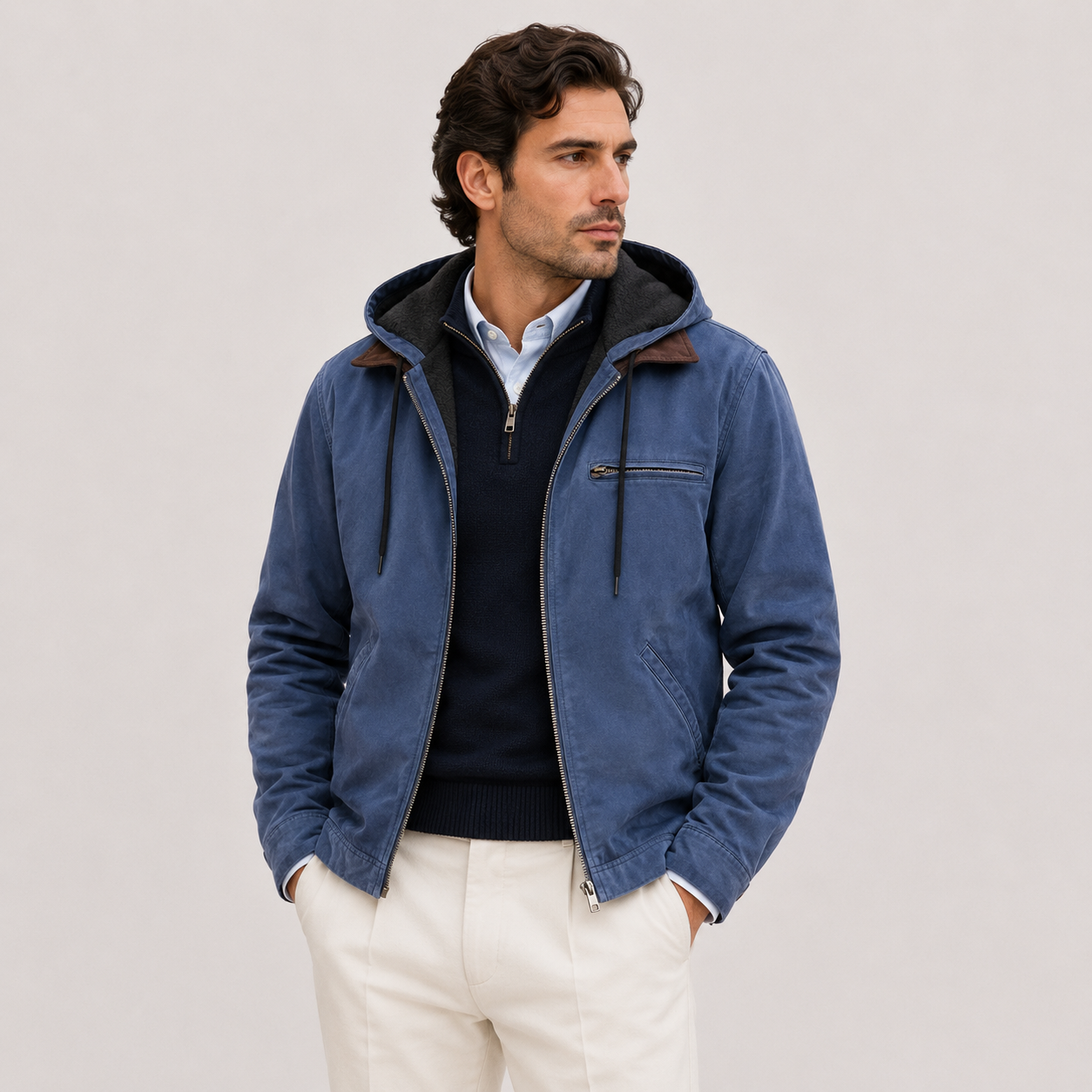 A man wearing a blue hooded zip-up jacket over a dark quarter-zip sweater and white trousers, standing against a plain light gray studio background.
