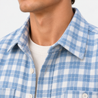 Close-up of a person wearing a light blue and white gingham plaid button-up shirt over a white ribbed knit undershirt, against a plain light background.