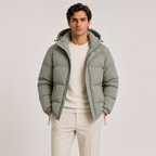 A man wearing a sage green hooded puffer jacket over a cream sweater and cream trousers, standing against a light gray studio background.