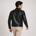 A man wearing a black leather jacket and beige trousers, photographed from behind against a light gray studio background.