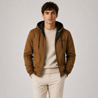 A man wearing a brown hooded jacket with a black-lined hood and a zippered chest pocket, layered over a cream sweater and cream trousers, standing against a light gray studio background.