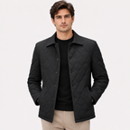 A man wearing a black diamond-quilted jacket over a black shirt and beige trousers, standing against a light gray studio background.