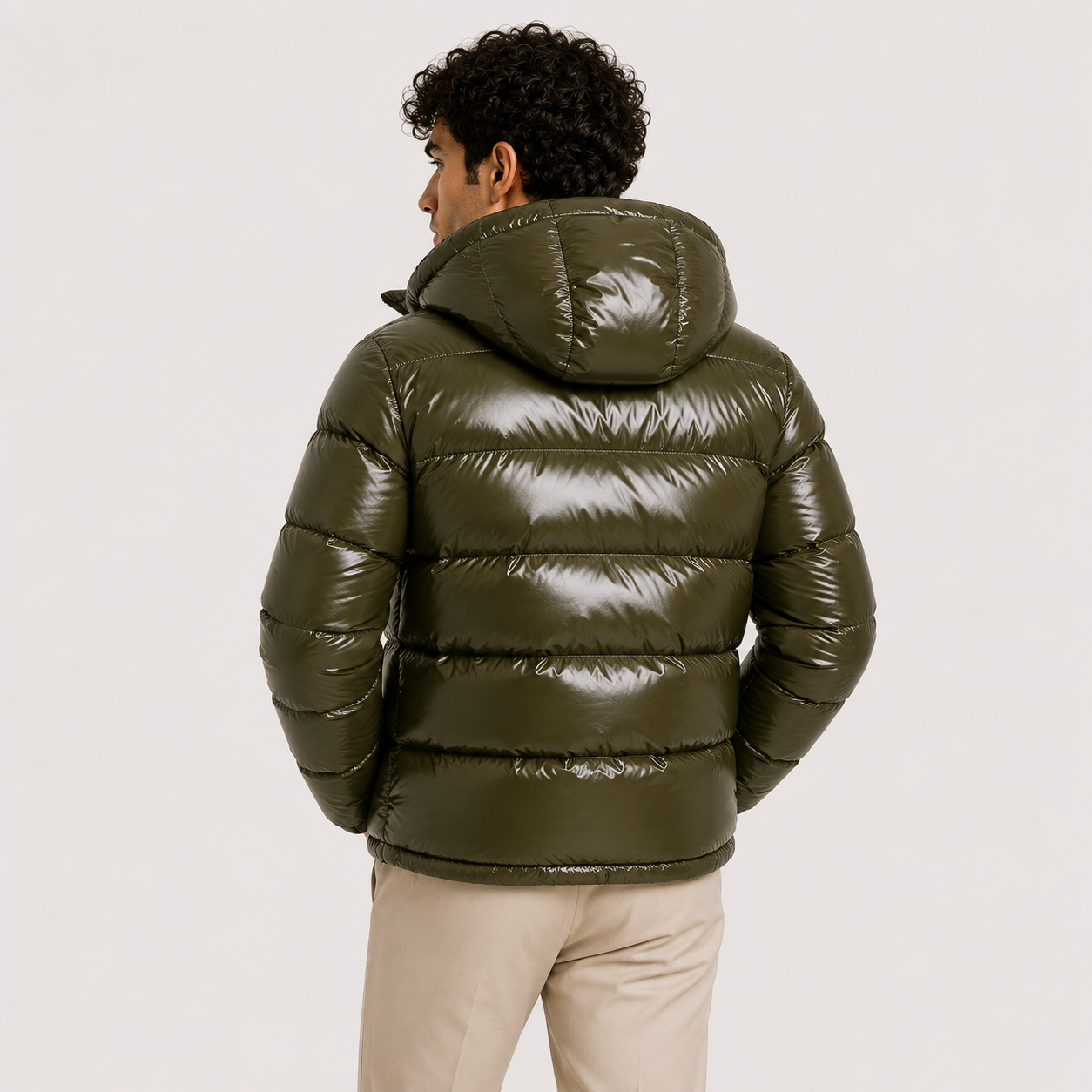 A man wearing a shiny olive green hooded puffer jacket, shown from the back against a plain light gray studio background.