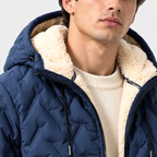 A close-up of a man wearing a navy blue quilted puffer jacket with a thick cream sherpa fleece lining on the hood and collar, layered over a cream ribbed knit sweater.