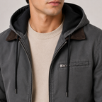 A close-up of a man wearing a dark gray hooded jacket with brown leather-look shoulder panels and black drawstrings, layered over a beige crewneck sweater.