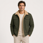 A man wearing an olive green corduroy jacket with a beige sherpa collar, layered over a cream sweater and beige trousers, photographed against a plain light gray studio background.