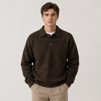 A man wearing a dark brown knit polo sweater over a white t-shirt and beige trousers, standing against a light gray studio background.
