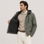 A man wearing an olive green hooded jacket with a black interior lining, styled over a beige sweater and white trousers, standing against a light gray studio background.