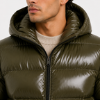 Close-up of a man wearing a shiny olive green puffer jacket with a high padded collar and black front zipper over a cream ribbed sweater.